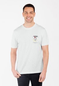 Hombre sonriendo, vistiendo una camiseta blanca con un pájaro y la palabra "ESPERANZA" impresa en el lado izquierdo del pecho, combinada con pantalones negros.
