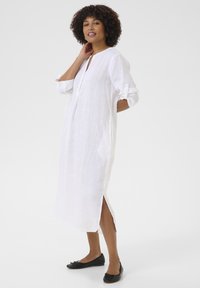 Abito bianco in lino con scollatura a V, maniche lunghe arrotolate ai polsi, spacchi laterali e vestibilità morbida, abbinato a ballerine nere.