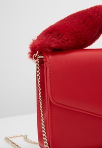 Červená kožená kabelka s akcentem z umělé kožešiny na vrchu, zlatým řetízkovým popruhem, kompaktním obdélníkovým tvarem a hladkým povrchem.