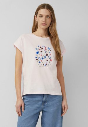 Kobieta z długimi włosami, ubrana w jasnoróżowy t-shirt z kolorowym kwiatowym nadrukiem i napisem „Kwiaty rozświetlają świat”, w połączeniu z niebieskimi dżinsami.