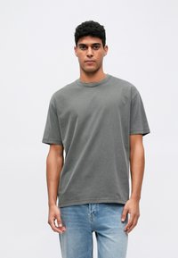 Šedé bavlnené tričko s krátkymi rukávmi a okrúhlym výstrihom, zobrazené na osobe, v kombinácii so svetlými modrými džínsami. Jednoduchý a neformálny dizajn.