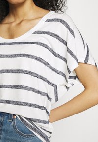 Mujer vestida con una camiseta blanca de manga corta con rayas horizontales negras y jeans de mezclilla azules, con una mano metida en el bolsillo del jeans.