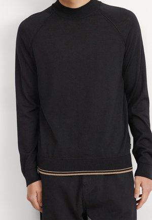 Man wearing a long-sleeve black sweater with a high neck and beige double stripe detail along the bottom hem.