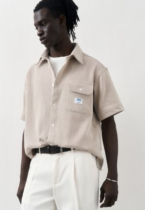 Mężczyzna w beżowej, fakturowanej koszuli na krótki rękaw z guzikami na białej koszulce, białe spodnie z czarnym paskiem i srebrna bransoletka na jednolitym tle.