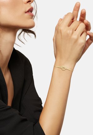 Kultainen rannekoru, jossa on sydämen muotoinen riipus, hienovarainen ketju ja pieniä pyöreitä korostuksia, kädessä tumman kankaan taustaa vasten.