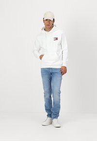Sudadera blanca con bolsillo canguro en la parte frontal y detalles del logo, combinada con jeans de mezclilla azul claro y zapatillas blancas. Un gorra beige completa el look.