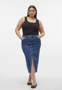 Fekete ujjatlan felső, amelyet magasderekú farmer szoknyával viselnek, elöl hasítékkal és oldalzsebekkel; a modell lapos cipőt hord.