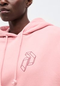 Primer plano de una persona que lleva una sudadera con capucha rosa con un diseño geométrico de un cubo abierto en el pecho.