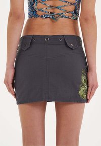 Grijs denimrok met een aansluitend ontwerp, voorzien van twee voorzakken, metalen hardware en gedetailleerde groene draakborduursels aan de zijkant.