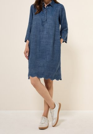 Kvinde iført blå knælængde kjole med hulbroderi på ærmer og kant, kombineret med hvide sneakers, stående med krydsede ben.