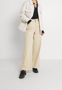 Beżowe spodnie o szerokich nogawkach połączone z pikowaną, jasnoszarą kurtką z kołnierzykiem i dwoma przednimi kieszeniami, noszone na czarnej koszuli.