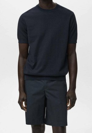 Homme portant une chemise bleu marine foncé unie à manches courtes avec un short bleu marine foncé assorti jusqu’aux genoux, debout devant un fond blanc.