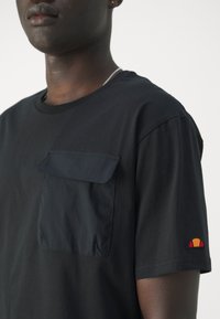 T-shirt noir à manches courtes avec une poche contrastante et un petit logo brodé en rouge, jaune et orange sur la manche gauche.