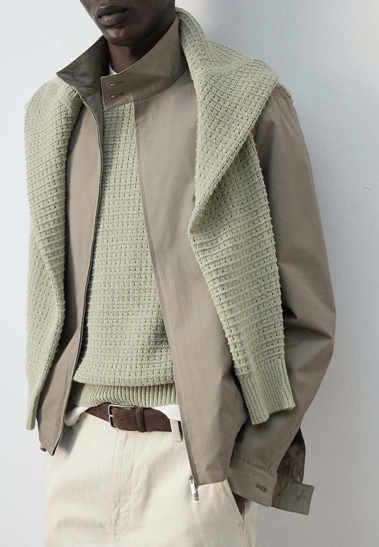 Personne portant une veste beige par-dessus un pull en maille vert clair avec une écharpe assortie en maille drapée sur les épaules, un pantalon blanc et une ceinture marron.