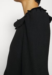 Svart texturerad tröja med volangkrage och puffärmar, som visar en mjuk tyg med subtila vertikala linjer.
