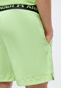 Άτομο που φοράει ανοιχτό πράσινο αθλητικό σορτς με μαύρη ζώνη μέσης Under Armour, το χέρι ακουμπά στην δεξιά τσέπη, σε απλό φόντο.