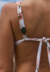 Top de bikini floral con tirantes ajustables, cierre de clip y espalda atada. Predominantemente blanco con acentos en naranja y verde.