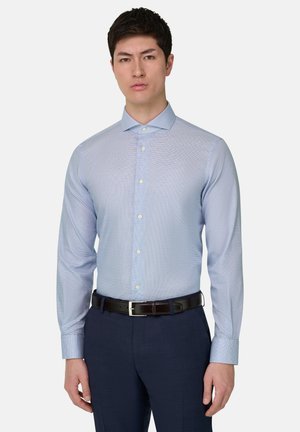 Mann trägt hellblaues Hemd mit dezentem Muster, dunkelblaue Hose und schwarzen Ledergürtel vor einfarbigem Hintergrund.