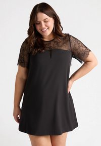 Schwarzes Kleid mit kurzen Spitzensäumen, einem Schlitz-Ausschnitt und einer lockeren, fließenden Silhouette. Glattes Material mit Spitzenverzierungen.