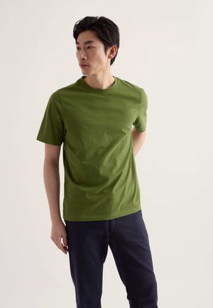 Homme debout regardant vers la gauche, portant un t-shirt vert uni à manches courtes et un pantalon foncé, sur un fond clair uni.