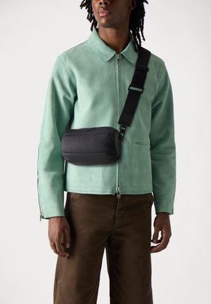 Borsa a tracolla nera imbottita con forma arrotondata, tracolla regolabile e chiusura con cerniera. Indossata sopra una giacca in suede verde e pantaloni marroni.