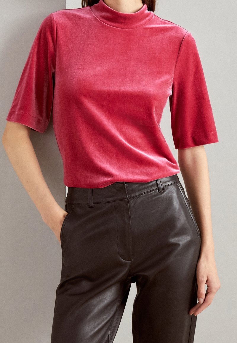 Haut à manches courtes et col montant en tissu doux, ressemblant à du velours, de couleur rose profond. Associé à un pantalon en cuir noir taille haute.