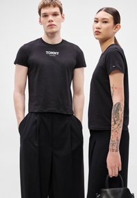 T-shirts pretas de algodão com o logotipo "TOMMY JEANS", gola redonda, corte slim. Os modelos vestem calças pretas, com um deles segurando uma pequena mala preta.