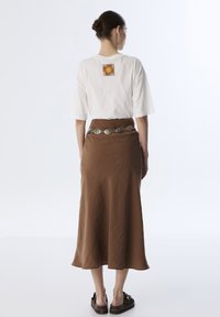 Camiseta blanca de manga corta con un gráfico en la espalda, combinada con una falda marrón de longitud media y un cinturón metálico con detalles decorativos.