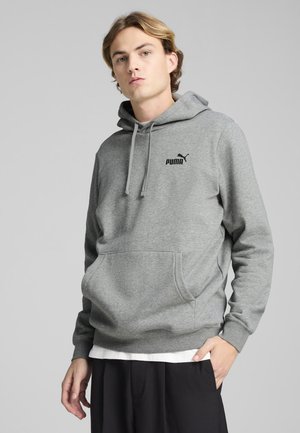 Joven vestido con una sudadera con capucha gris de Puma con bolsillo delantero y cordones, combinada con pantalones negros y una camiseta blanca interior.