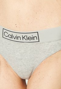 Tanga en coton gris avec une large ceinture élastique arborant le logo "Calvin Klein" en noir. Texture lisse, design minimal.