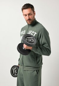 Manubrio nero tenuto in una mano, contrassegnato 12,5, con una presa in gomma. Il modello indossa una giacca verde con zip e lettering bianco e pantaloni abbinati.