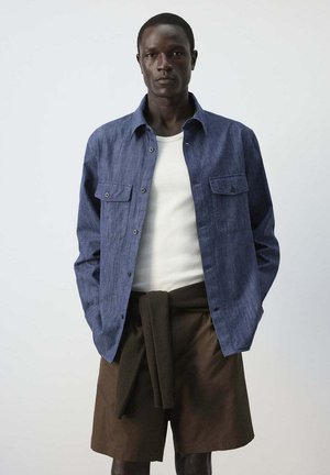 Joven con una camisa azul abotonada sobre una camiseta blanca, pantalones cortos marrones y un suéter oscuro atado alrededor de la cintura.