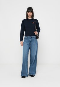 Granatowy sweter z dzianiny o prążkowanej teksturze, z małym logo. Jasnoniebieskie dżinsy o luźnym kroju z szerokimi nogawkami. Czarna torba na ramię.