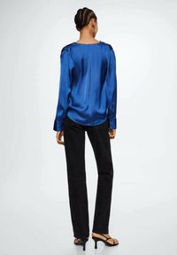 Blouse en satin bleu royal à manches longues avec détails aux épaules, associée à un pantalon noir et des talons noirs à brides, vue de dos.