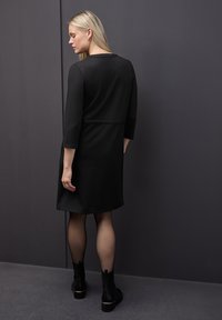 Schwarzes A-Linien-Kleid mit Dreiviertelärmeln, rundem Ausschnitt und einer Nahtdetails in der Taille. Getragen mit knöchellangen schwarzen Stiefeln.