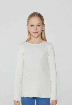 Haut à manches longues blanc en ribbed avec un col rond et un ourlet en scallop. Présente une texture douce, porté sur un jean bleu.