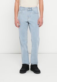 Személy világoskék egyenes szárú farmerben, fehér ingben és fekete tornacipőben, sima fehér háttér előtt állva.