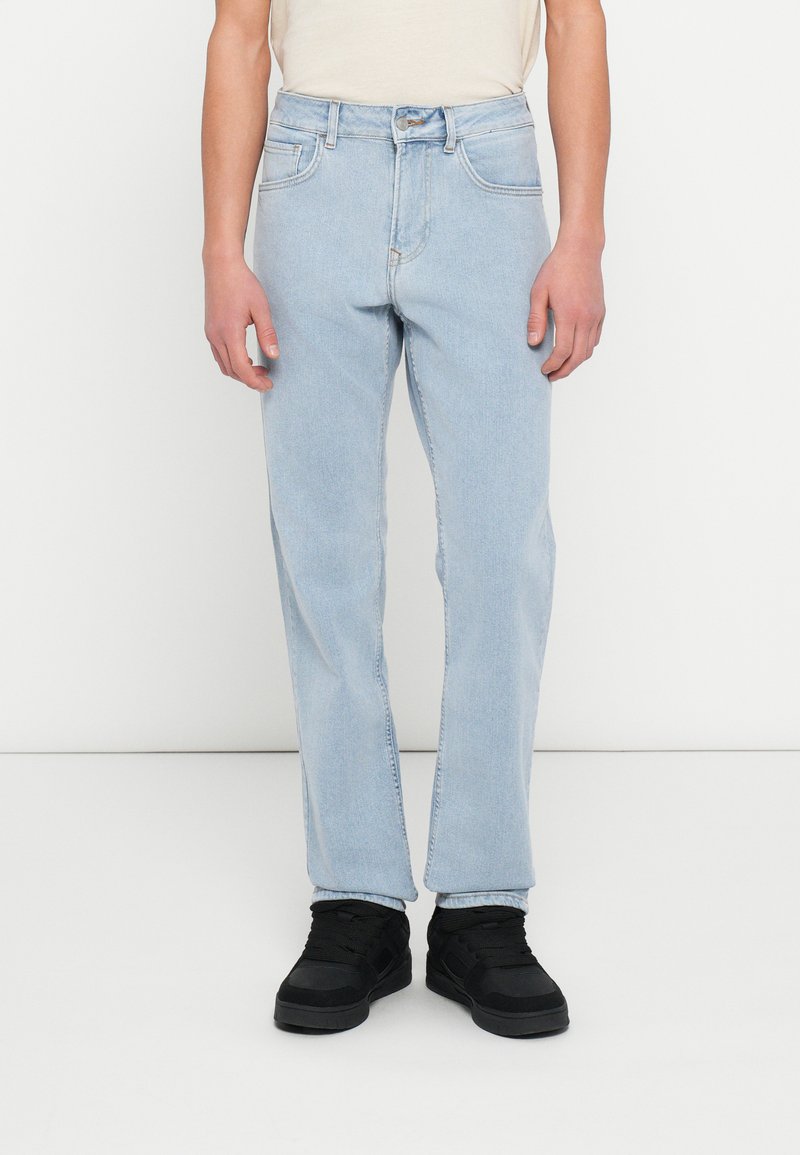 Személy világoskék egyenes szárú farmerben, fehér ingben és fekete tornacipőben, sima fehér háttér előtt állva.