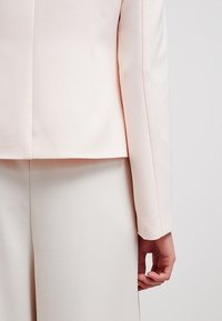 Jasnoróżowa marynarka o dopasowanym kroju, gładkiej fakturze, czystych liniach i wyraźnym fasonie, w zestawie z pasującymi jasnymi spodniami.