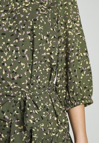 Robe verte avec des taches abstraites violettes, crème et noires, dotée de manches bouffantes trois-quarts et d'une ceinture en tissu nouée à la taille.