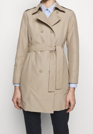 Beżowy, dwurzędowy płaszcz trencz z paskiem w talii, noszony na jasnoniebieskiej koszuli z kołnierzykiem i ciemnych spodniach, przycięty na wysokości ramion i do połowy ud.