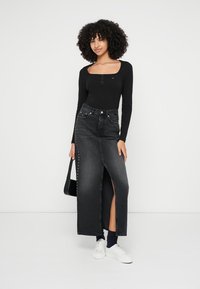 Top preto de manga comprida com botões, combinado com uma saia jeans escura de cintura alta, apresentando uma fenda frontal e um detalhe com tachas em um dos lados. Tênis brancos.