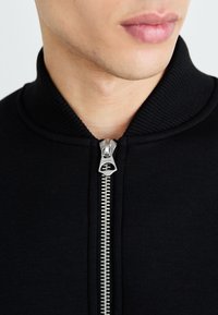 Veste noire zippée avec un col côtelé, présentant un détail de fermeture éclair métallique à l'avant. Texture de tissu douce et lisse.