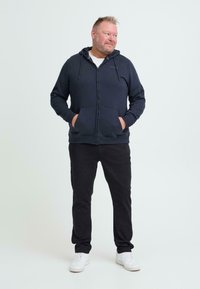 Felpa con zip blu navy, dotata di tasche frontali e cappuccio con cordino, abbinata a pantaloni neri e sneakers bianche, su uno sfondo chiaro.