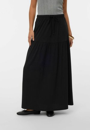 Kvinde iført en lang sort flæset nederdel med snøre i taljen, grå ærmeløs top, sorte flade sko og en brun armbånd om højre håndled.