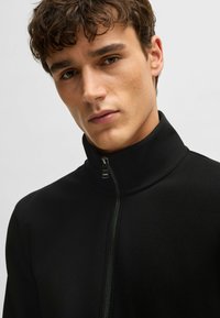 Zwarte zip-up jacket met hoge kraag, gemaakt van textuurstof. Beschikt over een ritssluiting aan de voorkant en een minimalistisch ontwerp.