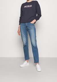 Sudadera marina con logotipo "HUGO" en blanco y rosa, combinada con jeans rectos de color azul claro y zapatillas blancas sobre un fondo neutro.