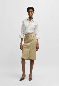 Light beige pencil skirt with a side slit and waistband detail, paired with a white striped button-up shirt. Model wears black pointed flats.