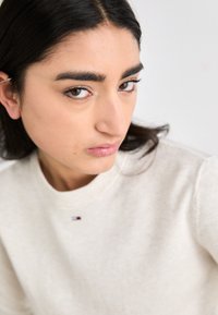 Pull crème avec un col rond et des poignets côtelés. Présente une petite étiquette de logo au niveau du col. Tissu doux et texturé.