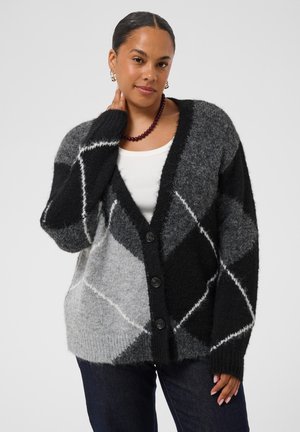Mulher a usar um cardigan preto e cinza estampado sobre uma camisa branca, com jeans escuros e um colar de contas roxo em fundo simples.